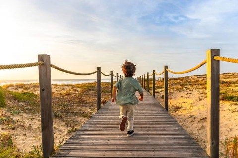 Little boy running fast along a wooden boardwalk towards a sandy beach and ocean, enjoying a summer