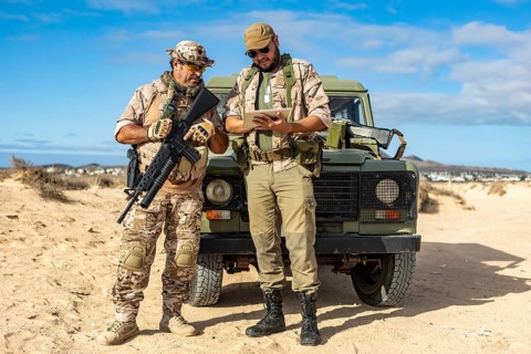 Two military soldiers in tactical gear stand in a desert landscape. One holds a rifle, while the
