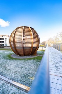 Large wooden sphere structure on grass, in the background modern buildings under a clear sky,