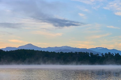 A still lake with morning mist and mountains in the background under a clear sky, Lake Mahinapua,