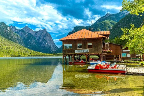 Pedal boats are docked near a restaurant on the shore of scenic lake dobbiaco with the dolomites in