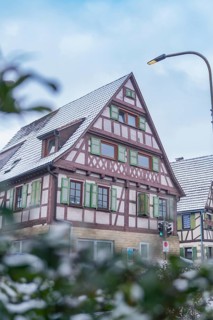 Snow-covered half-timbered house in a quiet street, surrounded by a few bushes and a lantern,