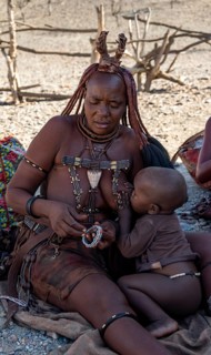 Traditional married Himba woman breastfeeding her baby, making bracelets, near Opuwo, Kaokoveld,