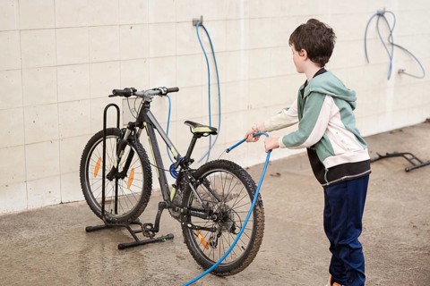 A young child attentively cleaning a bicycle with a hose in an outdoor space. The setting