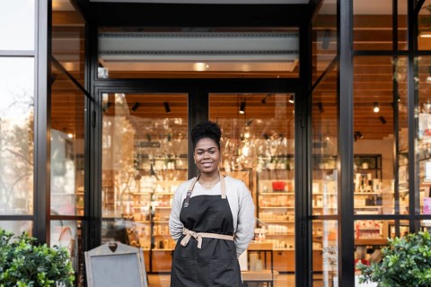 A friendly worker stands confidently in front of a vibrant cosmetics store, wearing an apron. The