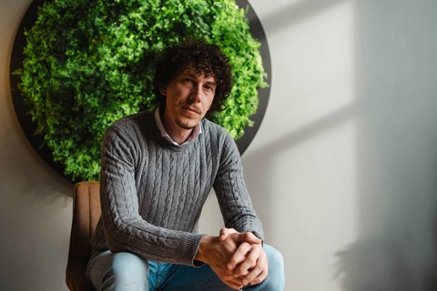Curly-haired young man sitting in front of a lush green wall. The setting emphasizes themes of