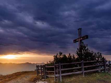 Summit cross on the Roßfeld under a dramatically overcast sky, the sun shines through a gap behind,