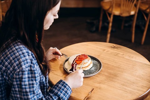 A woman in a plaid shirt sits at a wooden table, enjoying a plate of fluffy japanese pancakes