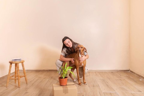 A young woman lovingly embraces her dog in a sparsely furnished room, surrounded by a stool, a