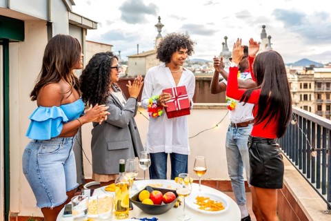 Multi-ethnic young chic friends applauding while man receiving a birthday gift during rooftop party