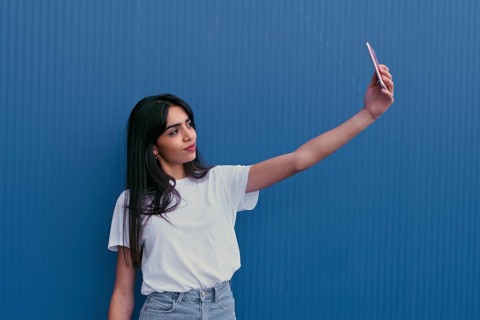 A young woman in a white shirt takes a selfie against a vibrant blue wall. Her confident expression