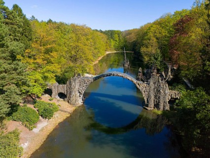 Round bridge over a quiet lake in a green forest with autumn trees and clear blue sky, aerial view,