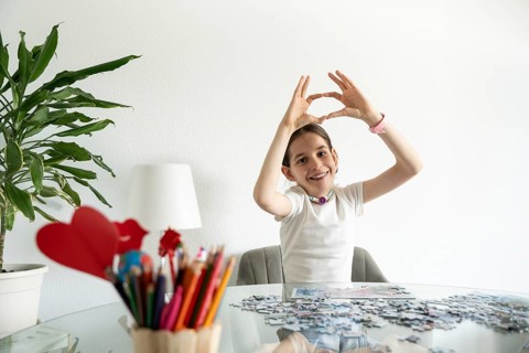 Smiling disabled girl with tracheostomy sits at a table with a puzzle. Colored pencils and heart