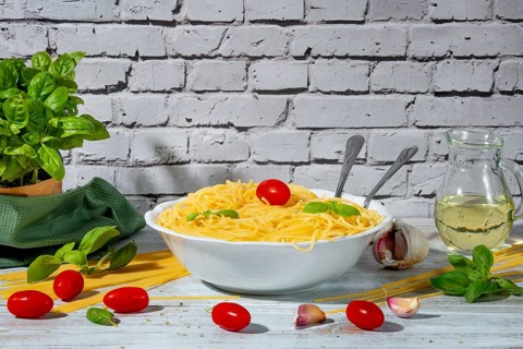 Bowl of cooked spaghetti surrounded by tomatoes, basil, olive oil and garlic on a table