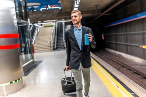 Young businessman walking through a bustling subway station, carrying a suitcase and sipping coffee