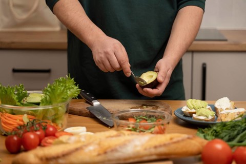 A person skillfully prepares an avocado, surrounded by fresh vegetables and bread on a kitchen