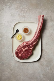 Top-down view of a raw tomahawk steak presented on a ceramic tray, accompanied by a small bowl of