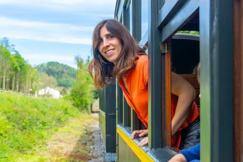 A woman is sitting in a train car with her head out the window. She is smiling and looking out at