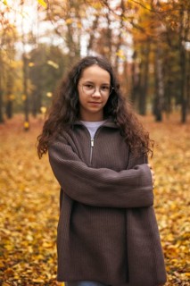 A girl in a cozy brown sweater stands amid a forest filled with vibrant autumn leaves. Her serene