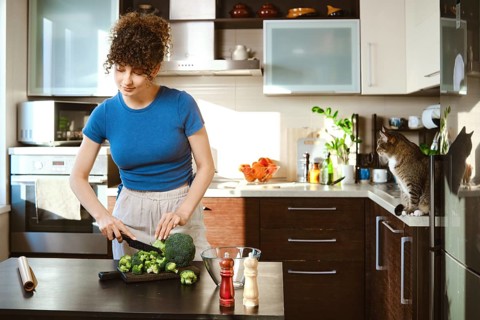 Woman is cutting broccoli on black cutting board in bright kitchen. Cat sits on the counter nearby
