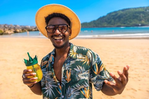 Young man with straw hat and hawaiian shirt smiling and holding pineapple cocktail on a sunny beach