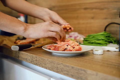 Hands are shredding cooked pink salmon on plate in kitchen. Fresh vegetables like green beans and