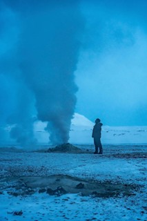 A man stands in the snow looking at a large plume of smoke. The sky is a deep blue color, and the