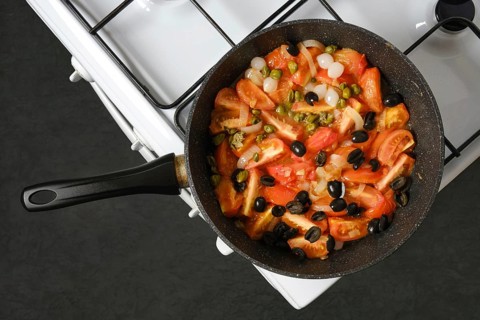 A pot sits on a stove with chopped tomatoes, pearl onions, olives and capers