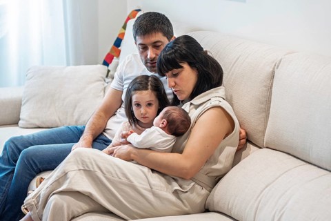 A young family sits closely together on a cozy couch, with a joyful big sister gazing at her