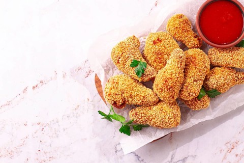 A wooden board holds several golden chicken drumsticks arranged neatly. A small bowl of red dipping