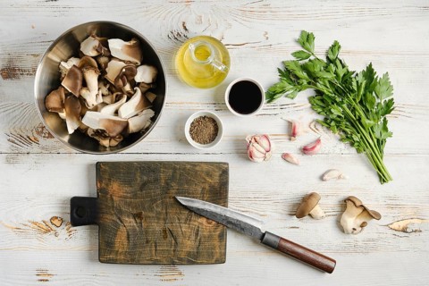 Oyster mushrooms in bowl alongside cooking oil, garlic cloves, and fresh herbs. Cutting board with