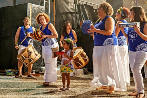 Women with instruments laugh while a little girl is integrated into the drum line, a band plays and