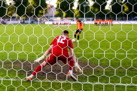 View through the net of a soccer goalkeeper in a red kit preparing to defend a penalty kick during