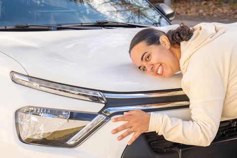 Smiling woman playfully resting her cheek on car hood while hugging it, close-up shot expressing