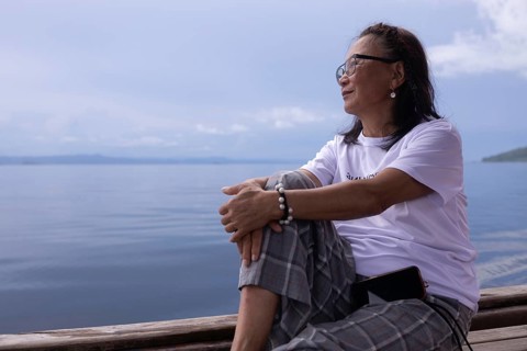 A mature woman sits contentedly on a wooden deck on Kri Island, looking out over the calm waters of