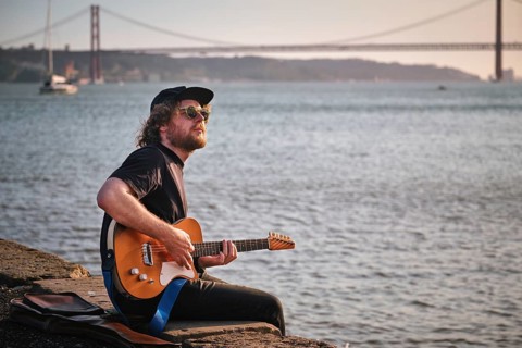 Hipster street musician in black playing electric guitar in the street sitting on pier embankment