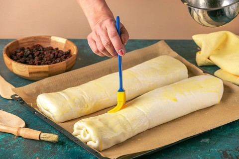 Hand spread strudel with egg on baking tray next to bowl with raisins