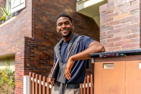 African young businessman leaning in a residential house entrance carrying a laptop and smiling at
