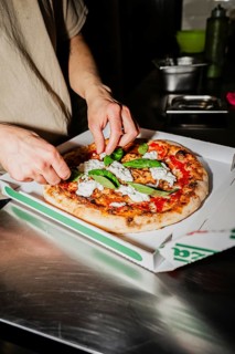 A chef adds fresh basil to a gourmet pizza with ricotta cheese in a cardboard box. The kitchen