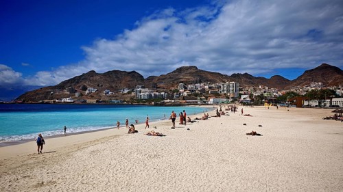 Sunny sandy beach 'Praia da Laginha' with holidaymakers and blue sea against a mountain backdrop,