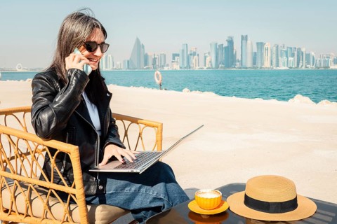 Happy business woman drinking coffee using laptop and smartphone chat. West Bay skyline background