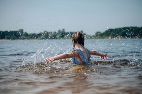 A young girl with braided hair plays joyfully in a lake, splashing water under a clear blue sky.