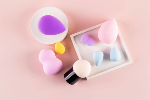 Top view of assorted makeup sponges, including a beauty blender, arranged on a pink background,