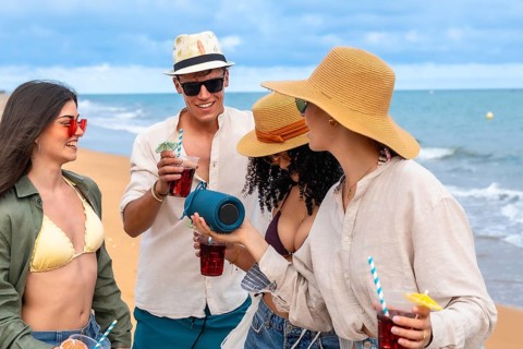 Young friends chilling on a sunny beach with colorful cocktails and a portable speaker, enjoying