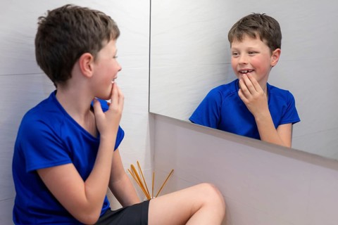 A young boy in a blue shirt smiles at his reflection, touching the gap in his teeth where a milk