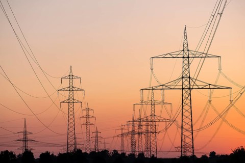 Silhouette of several high-voltage power lines and electricity pylons against a glowing orange and