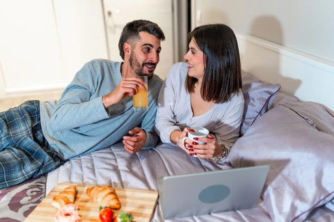 Young couple lying in bed, having breakfast and watching something on their laptop, spending