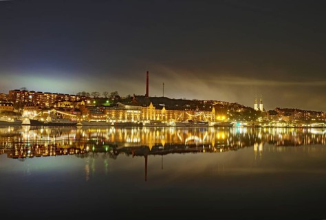 Illuminated buildings reflected in calm water, surrounded by light haze at night, faint aurora