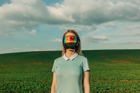 A woman stands in a vast green field wearing a futuristic rainbow visor. The vibrant colors of the