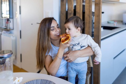 Mother sharing a slice of pizza with her toddler son while sitting together at the kitchen table,